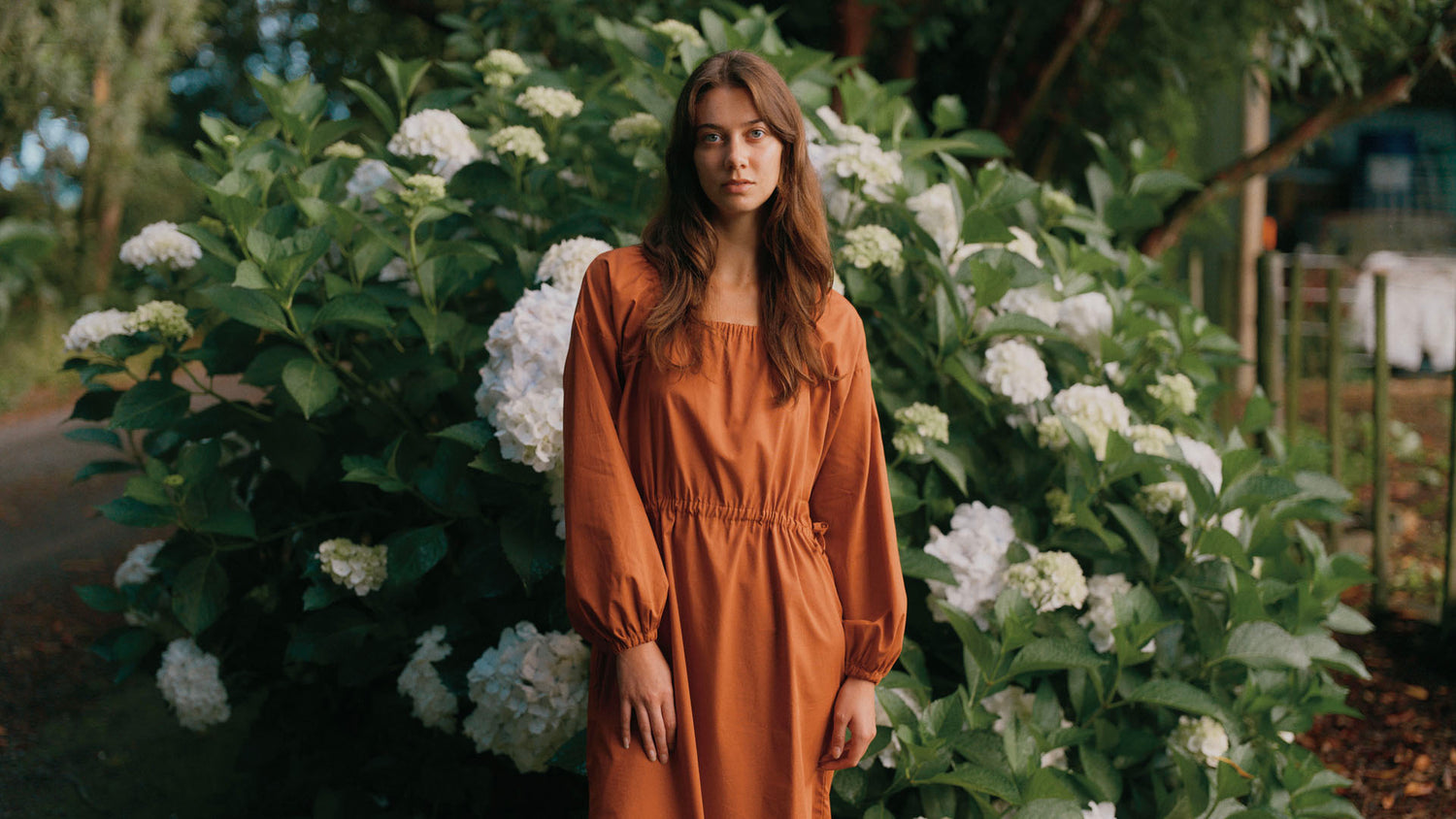 Women wearing a Burnt Sienna dress, standing in front of a bush with white flowers