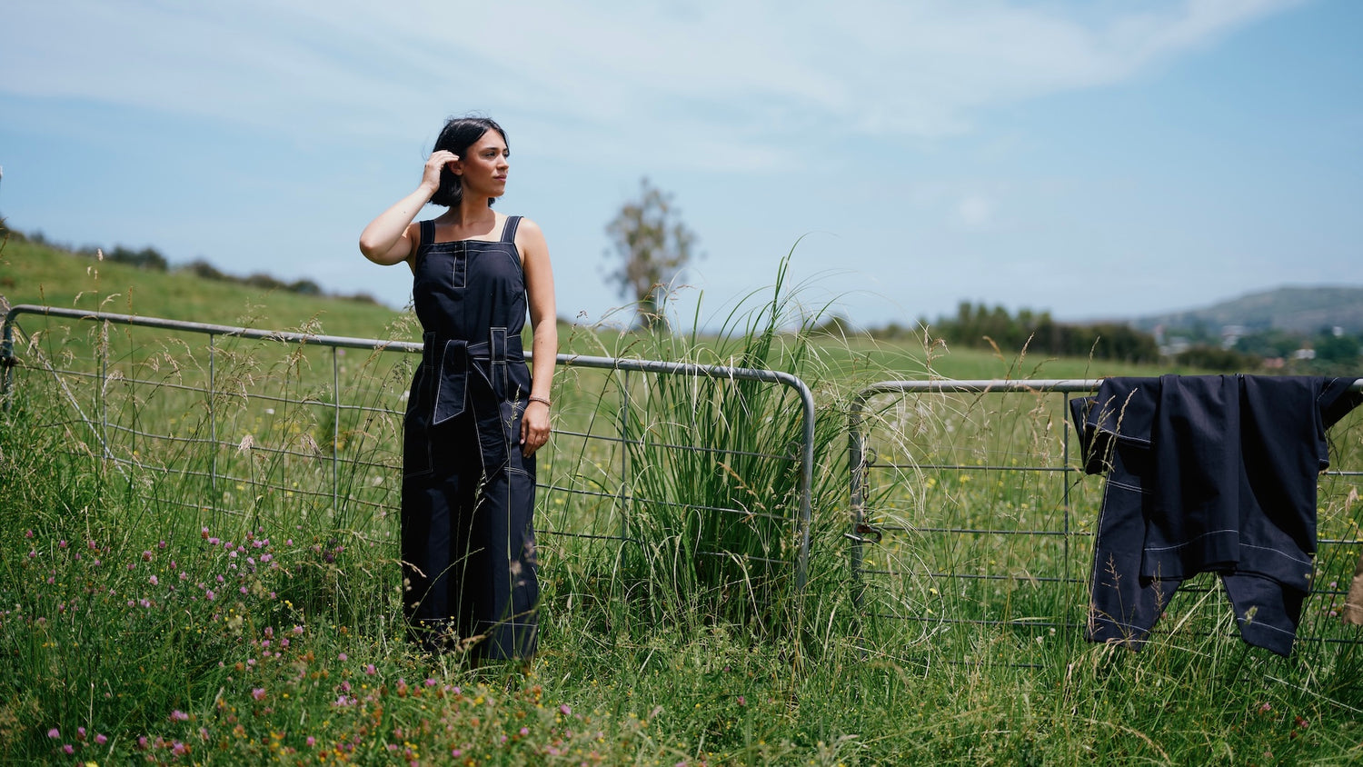 A woman stands in a grassy field wearing a dark indigo sleeveless jumpsuit with wide legs and a tie waist. A matching dark garment hangs on a wire fence to the right.
